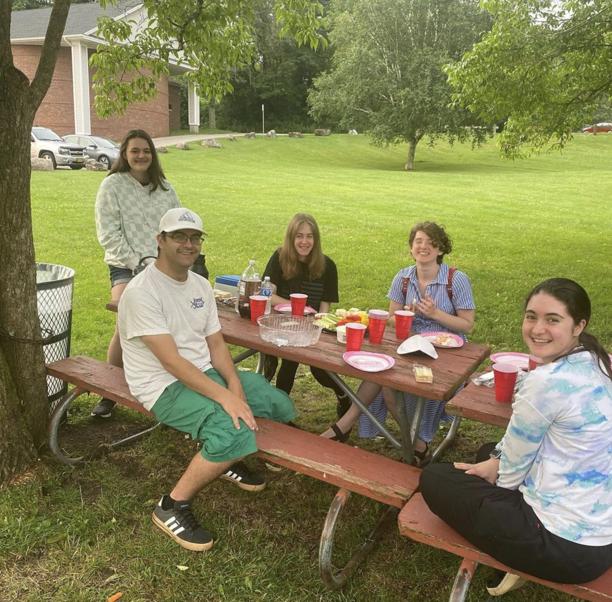 A group of young adults sit around a picnic table smiling at the camera.