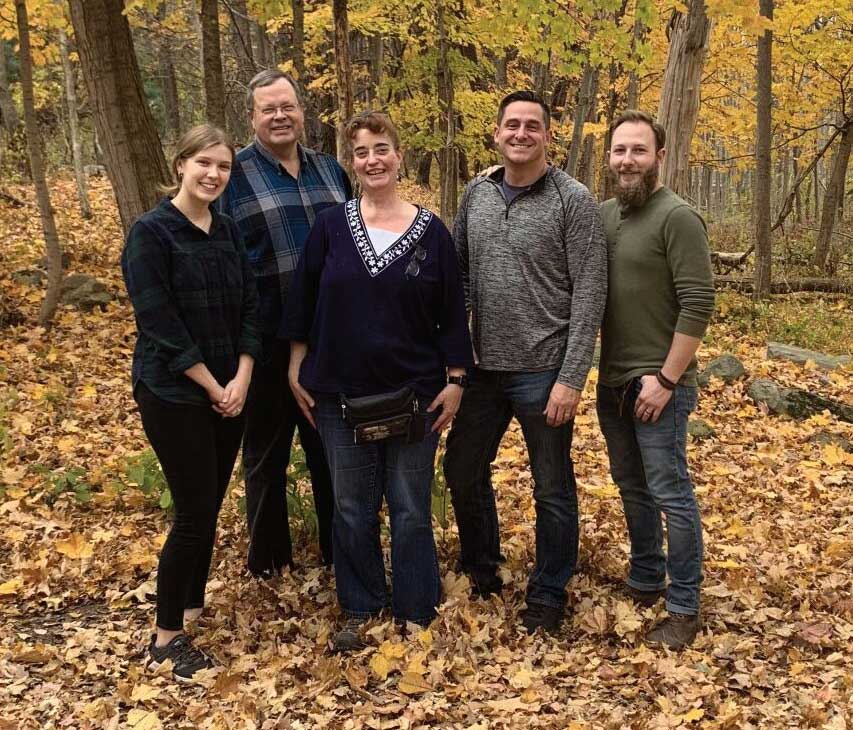 The staff of five from Pittsford Community Church stand in a group in autumn woods, smiling at the camera.